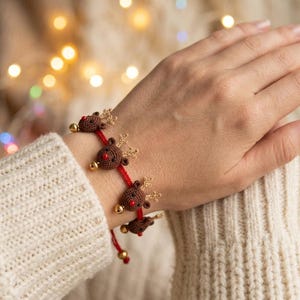 May include: A red cord bracelet featuring multiple small, crocheted brown reindeer charms with red noses, tan antlers, and tiny gold-toned metallic bells attached between each reindeer figure along the length of the adjustable string band.