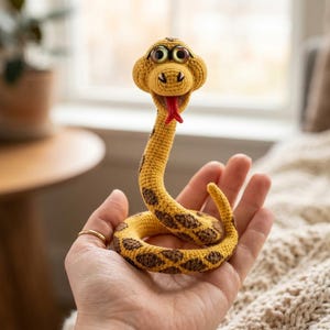 May include: A yellow crocheted snake toy with brown diamond-shaped spots, large round eyes, and a red tongue. The snake is coiled in a circular shape with its head raised upright, featuring a textured yarn surface and detailed stitching throughout.