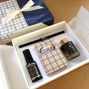 May include: A gift set in a white box containing a black spray bottle labeled Aroma Spray, a rectangular box with a beige and blue plaid pattern featuring small white hearts, and a glass diffuser bottle labeled Diffused Velora 50ml / 1.7oz.