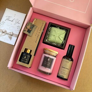 May include: A pink gift box containing a Nature's Embrace diffuser set with a black bottle, a square green wrapped item, a pink candle in a glass jar with a wooden lid, and an amber spray bottle labelled Aroma Spray, plus a Thank You gift card.