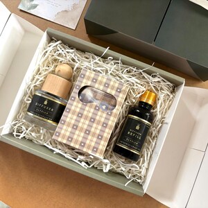 May include: A gift box containing a glass diffuser with a wooden cap, a small brown plaid box with a heart pattern, and a dark glass bottle labeled Diffuser Refill Elysian. The items are nestled in white paper shred filler inside a green box.