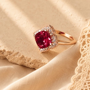 May include: A rose gold ring featuring a large, cushion-cut deep red gemstone at the center. The stone is surrounded by a halo of small, clear, sparkling white stones set in a scalloped, floral-inspired metal frame that matches the band.