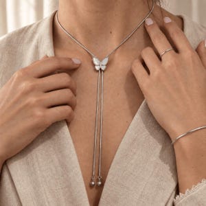 May include: A silver-toned lariat necklace featuring a central butterfly pendant with white inlay. The necklace has a thin chain that extends downward from the butterfly, ending in two small silver-toned spherical beads at the bottom of the strands.
