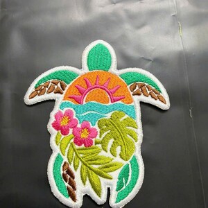 May include: An embroidered patch shaped like a sea turtle with a white border. The shell features a vibrant orange sun with pink rays, a light blue ocean, two bright pink hibiscus flowers, and green tropical monstera and fern leaves.