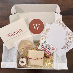 May include: A gift box containing a Warmly card, a gold-toned leaf-shaped ornament, a small wooden candle holder with a white candle, a red string bracelet with a floral bead, and a bag of organic cranberry health mix on shredded paper.