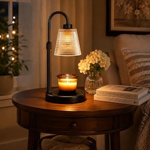 May include: A black metal candle warmer lamp with a curved arm and a clear, textured glass shade. The lamp features a round base holding a small amber glass jar candle that is illuminated from above, casting a warm glow on the surrounding area.