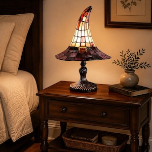 May include: A stained glass table lamp shaped like a pointed hat with a wide brim. The lamp features a color palette of warm orange, cream, and deep purple glass panels, accented by a small buckle detail. It rests on a dark, ornate metal base.