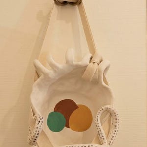 Peut inclure: Un bol mural en céramique blanche orné de trois cercles peints à la main en vert, marron et jaune. Le bol est suspendu par une sangle en tissu beige et décoré de deux bras en céramique blanche tachetée en forme de tentacules.