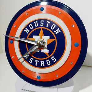 May include: A blue and orange clock with a white face and the words "Houston Astros" in a navy blue circle. The clock has silver hands and blue accents.