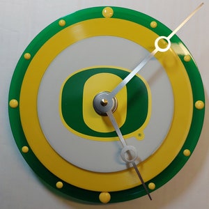 May include: A green and yellow circular clock with a white face and a silver metal 'O' logo. The clock has a green outer ring with yellow dots and a yellow inner ring with green dots.