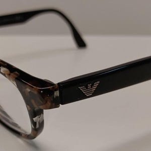 May include: Eyeglasses featuring a tortoiseshell pattern on the front frames and solid black temples. A silver-toned eagle logo is prominently displayed on the side of the temple near the hinge. The frames are constructed with a classic rectangular shape.