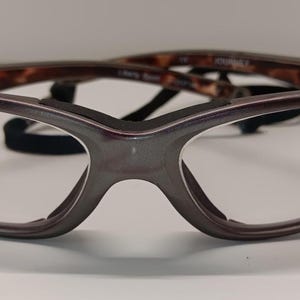 May include: A pair of eyeglasses with a dark brown frame and clear lenses. The temples have a tortoiseshell pattern. A black strap is attached to the frame. The glasses are designed for vision correction.