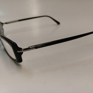May include: Black Prada eyeglasses with rectangular frames. The temples are black with silver accents and the Prada logo. The glasses are set against a white background.