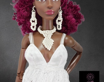 May include: A doll with dark skin and curly magenta hair wearing a white lace dress, a white beaded choker necklace, and matching white beaded drop earrings. The doll has brown eyes and pink lips. A black square logo with a diamond icon and the text LoReeby Lane is visible in the bottom right corner.