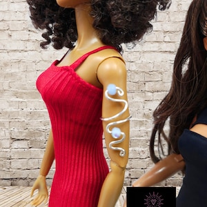 May include: A silver-toned wire arm cuff featuring a swirling, serpentine design accented with two small, pale blue beads. The accessory is worn on the upper arm of a doll dressed in a sleeveless, ribbed red dress with thin shoulder straps.