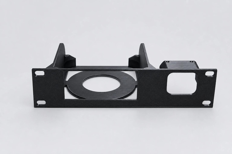 May include: A black rack-mount panel featuring a circular cutout in the center, a square cutout on the right side, and four mounting holes at the corners. The structure includes two vertical support fins extending backward from the main faceplate.