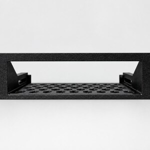 May include: A black metal rack-mount shelf featuring a rectangular front panel with four mounting holes at the corners. The shelf base has a perforated pattern of hexagonal cutouts for ventilation and two side support brackets.