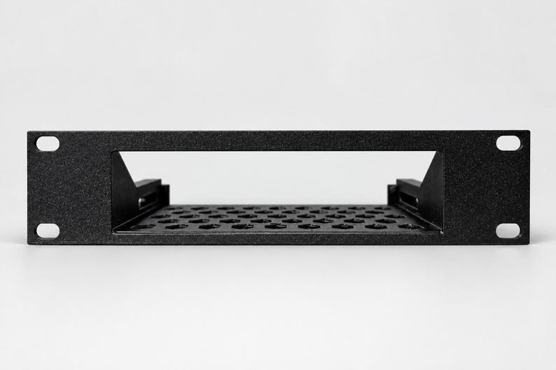 May include: A black metal rack-mount shelf featuring a rectangular front panel with four mounting holes at the corners. The shelf base has a perforated pattern of hexagonal cutouts for ventilation and two side support brackets.