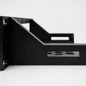 May include: A black metal rack-mount shelf featuring a flat base with a perforated pattern of circular holes, side support brackets with rectangular cutouts, and a vertical front panel with two circular mounting holes on each side.