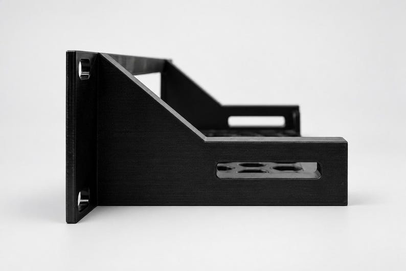 May include: A black metal rack-mount shelf featuring a flat base with a perforated pattern of circular holes, side support brackets with rectangular cutouts, and a vertical front panel with two circular mounting holes on each side.