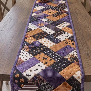 May include: A rectangular quilted table runner featuring a patchwork design of various Halloween-themed fabrics in shades of orange, purple, black, and white. The pattern includes motifs of pumpkins, bats, spider webs, cats, and haunted houses.