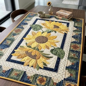 May include: A rectangular quilted table runner featuring three large yellow sunflowers with brown centers and green leaves in the center. The border consists of a log cabin quilt pattern in shades of blue, green, and brown with a yellow fabric edge.