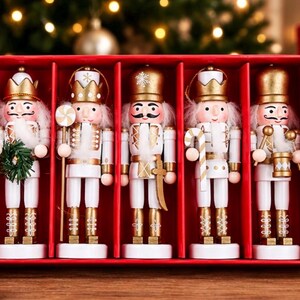 Op de afbeelding: Een set van vijf notenkraker-ornamenten in een rode doos. Elke notenkraker draagt een gouden kroon of hoed en een wit uniform met gouden accenten. Ze houden verschillende voorwerpen vast, waaronder een groene krans, een lolly, een zwaard, een zuurstok en een trommel.