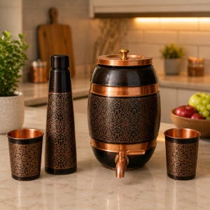 Puede incluir: Un conjunto que consta de un dispensador de agua en forma de barril con grifo de cobre, una botella alta y dos vasos a juego. Cada artículo presenta un acabado marrón oscuro con un intrincado patrón floral de color cobre y detalles metálicos.