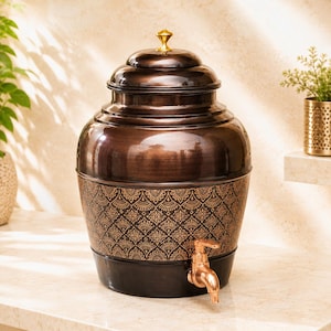 May include: A large, rounded copper-toned water dispenser with a dark, brushed metallic finish. The lower section features an intricate, repeating geometric pattern in a lighter copper hue. It includes a matching lid with a small gold-coloured knob.