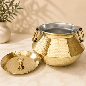 May include: A gold-colored hammered metal pot with a matching lid and two circular side handles. The pot features a wide, rounded body with a textured exterior and a smooth, silver-toned interior lining, presented with the lid placed beside it.