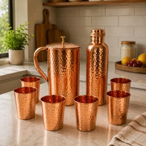 May include: A set of hammered copper drinkware consisting of a large pitcher with a lid and handle, a tall water bottle with a cap, and five matching tumblers. Each piece features a textured, dimpled surface with a warm, metallic copper finish.