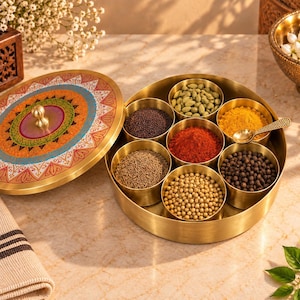 Puede incluir: Un especiero redondo de latón con siete recipientes pequeños extraíbles llenos de diversas especias, incluyendo cardamomo verde, semillas de mostaza, cúrcuma, chile en polvo, comino, semillas de cilantro y granos de pimienta negra, con tapa.