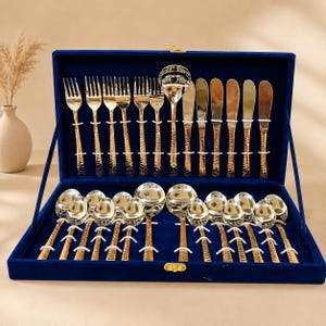 Puede incluir: Un juego de cubiertos con mangos dorados de acabado martillado y cabezas plateadas pulidas. La colección incluye tenedores, cuchillos y cucharas, dispuestos ordenadamente dentro de un estuche de presentación forrado en terciopelo azul.