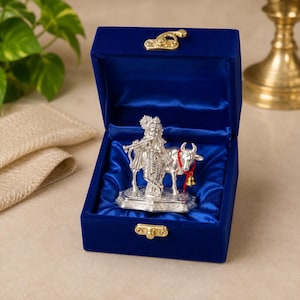 May include: A silver-toned figurine of a deity playing a flute while standing beside a cow, presented inside a deep blue velvet-lined box with a gold-coloured metal latch. The cow wears a red string with a small gold-coloured bell around its neck.