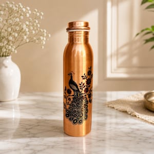 Op de afbeelding: Een cilindervormige koperen waterfles met een donkere, gedetailleerde grafische print van een pauw op een bloeiende tak. De fles heeft een gladde, metalen afwerking en een bijpassende schroefdop in een warme, gepolijste koperkleur.
