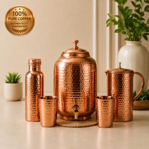 Puede incluir: Un juego de vajilla de cobre martillado que incluye un gran dispensador de agua con grifo, una botella alta, una jarra con asa y dos vasos a juego. Un sello dorado en la esquina dice 100% PURE COPPER AUTHENTICITY GUARANTEED.