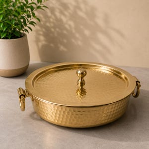 May include: A round, gold-colored metal serving dish with a hammered texture. It features a matching lid with a central knob handle and two side handles shaped like rings. The polished finish reflects light across the dimpled surface.