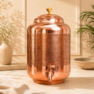 Op de afbeelding: Een cilindervormige koperen waterdispenser met een textuurrijke, gehamerde band rond het midden. De houder heeft een deksel van gepolijst koper met een kleine, ronde goudkleurige knop bovenop en een bijpassende koperen kraan aan de onderkant.