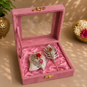 Puede incluir: Un joyero de terciopelo rosa con tapa transparente que contiene dos objetos decorativos plateados. Uno tiene forma de hoja con una rosa roja y una gema rosa, y el otro es un broche en forma de pluma con un detalle central rojo y dorado.