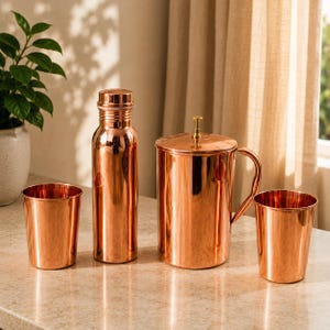 May include: A polished copper drinkware set consisting of a tall water bottle with a screw-top lid, a cylindrical pitcher with a handle and a small knob on the lid, and two matching drinking tumblers, all featuring a smooth, reflective finish.
