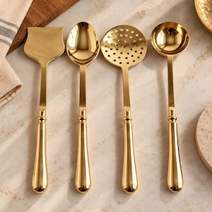 Könnte beinhalten: Ein Set aus vier goldfarbenen Küchenutensilien mit polierter Oberfläche. Die Kollektion umfasst einen breiten flachen Pfannenwender, einen ovalen Löffel, einen runden Schaumlöffel mit vielen Löchern und eine tiefe Schöpfkelle mit langem Griff.