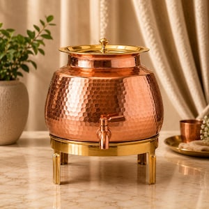 Pode incluir: Um dispensador de bebidas de cobre martelado com uma tampa dourada polida e um suporte de metal dourado a condizer. O dispensador possui uma torneira central de cobre para servir líquidos e assenta numa base circular com quatro pernas.