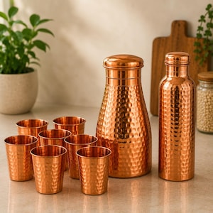 Puede incluir: Un juego de recipientes de cobre martillado que consta de seis vasos pequeños, una jarra de boca ancha con tapa y una botella de agua alta. Cada pieza presenta un acabado de cobre metálico reflectante con una superficie texturizada y abollada.