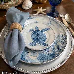 May include: A silver charger plate with a beaded rim, topped with a dinner plate and a smaller salad plate featuring a blue sea turtle illustration. A blue cloth napkin is secured with a jute ring, accompanied by a blue glass and silverware.