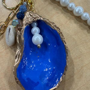 May include: A decorative keychain featuring a gold-toned oyster shell with a vibrant blue interior. The shell is accented with a dangling white pearl, a small seashell charm, and a gold chain. A strand of white beads is attached to the gold ring.