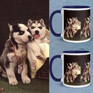 May include: Two ceramic mugs with a dark blue handle and rim, featuring a photographic print of two husky puppies. One puppy wears a red toy stethoscope, while the other sits in a basket with its tongue out. The design is repeated on both mugs.