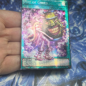 Può includere: Una carta collezionabile olografica di Yu-Gi-Oh! intitolata Pot of Greed, con un'illustrazione stilizzata di un personaggio biondo in un completo rosa accanto alla creatura Pot of Greed. La carta ha uno sfondo turchese e viola con cuori.