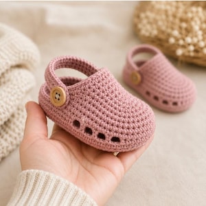 Könnte beinhalten: Ein Paar gehäkelte Babyschuhe in Altrosa mit abgerundeter Zehenpartie, einem schwenkbaren Fersenriemen, der auf beiden Seiten mit einem hellbraunen Holzknopf befestigt ist, und einer Reihe kleiner rechteckiger Ausschnitte am Sohlenrand.