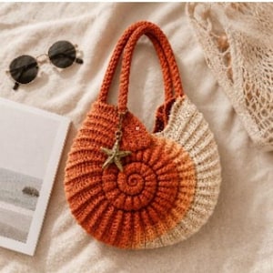 May include: A round, spiral-shaped crochet handbag featuring an ombre colour gradient transitioning from deep orange to cream. The bag includes two matching crochet handles and a metallic starfish charm attached to the top of the spiral design.