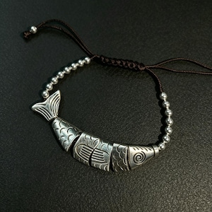May include: A bracelet featuring a central silver-toned fish charm composed of three articulated segments with etched patterns. The fish is flanked by small silver-toned beads on a dark brown adjustable cord with a sliding knot closure mechanism.